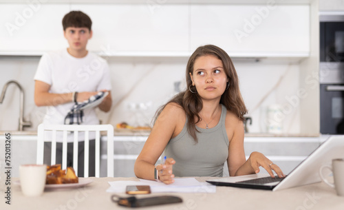 Annoyed young girl doing business on notebook while guy is brawling with her at the kitchen table