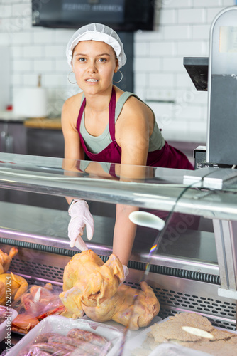 In butcher shop, young female salesperson in gloves shows damp dressed chicken. Seller takes pan-ready hen out of refrigerator window photo