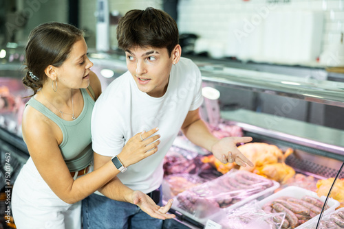 Attentive young man and woman purchasers choosing sausages from closed-type showcase in butcher shop