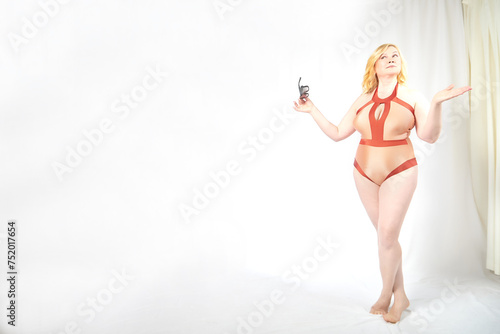 Funny cute mature adult woman with blonde hair and sunglasses in a swimsuit on a white background. Fat middle-aged lady poses in underwear in studio. The concept of body positivity