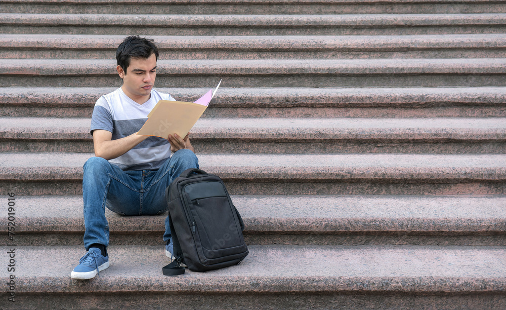 A male student sitting on the school stairs reading a document
