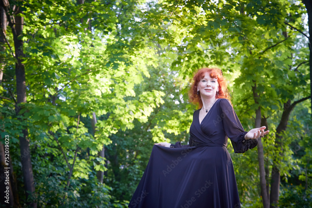 Elegant senior mature Woman dancing in Black Dress in green park. Woman with red hair posing on nature landscape Elegant senior mature Woman dancing in Black Dress in green park. Woman with red hair posing on nature landscape