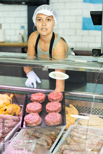 Positive young saleswoman standing behind counter demonstrating a tray with burger patties in butcher shop