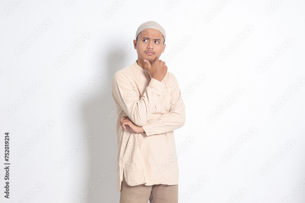 Portrait of confused Asian muslim man in koko shirt with skullcap thinking about question with hand on chin. Education concept. Isolated image on white background Portrait of confused Asian muslim man in koko shirt with skullcap thinking about question with hand on chin. Education concept. Isolated image on white background