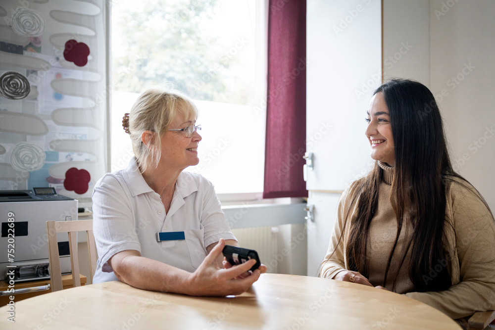 Smiling female doctor showing glucometer to woman at table in clinic Smiling female doctor showing glucometer to woman at table in clinic