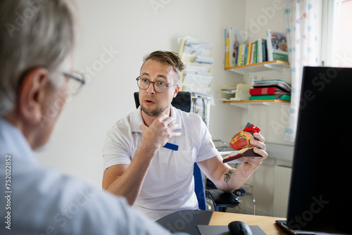 Male doctor with heart model explaining to elderly man in clinic photo