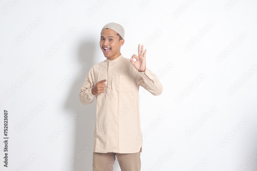 Portrait of young attractive Asian muslim man in koko shirt with skullcap showing ok hand gesture and smiling looking at camera. Approval concept. Isolated image on white background Portrait of young attractive Asian muslim man in koko shirt with skullcap showing ok hand gesture and smiling looking at camera. Approval concept. Isolated image on white background