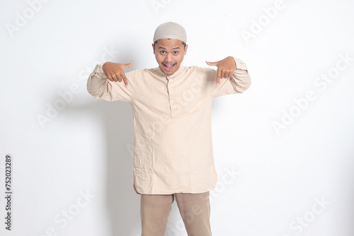 Portrait of shocked Asian muslim man in koko shirt with skullcap showing product and pointing with his hand and finger down. Advertising concept. Isolated image on white background