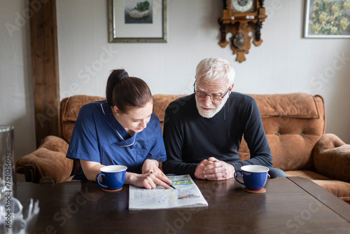 Female caregiver teaching senior man to solve crossword puzzle in living room at home