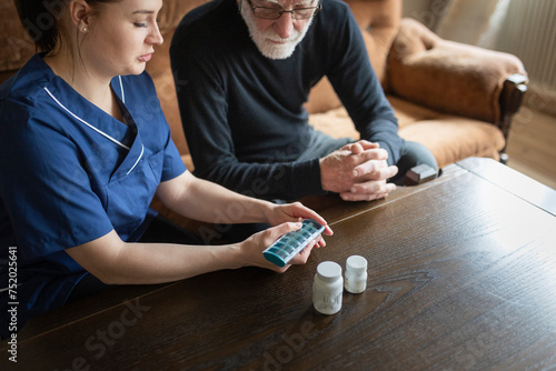 Female nurse with pill box giving advice to elderly man at table in living room photo
