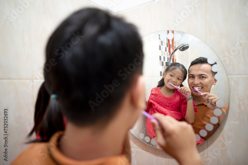 Indonesian father with daughter brush their teeth in bathroom looking through the oval mirror at home.
