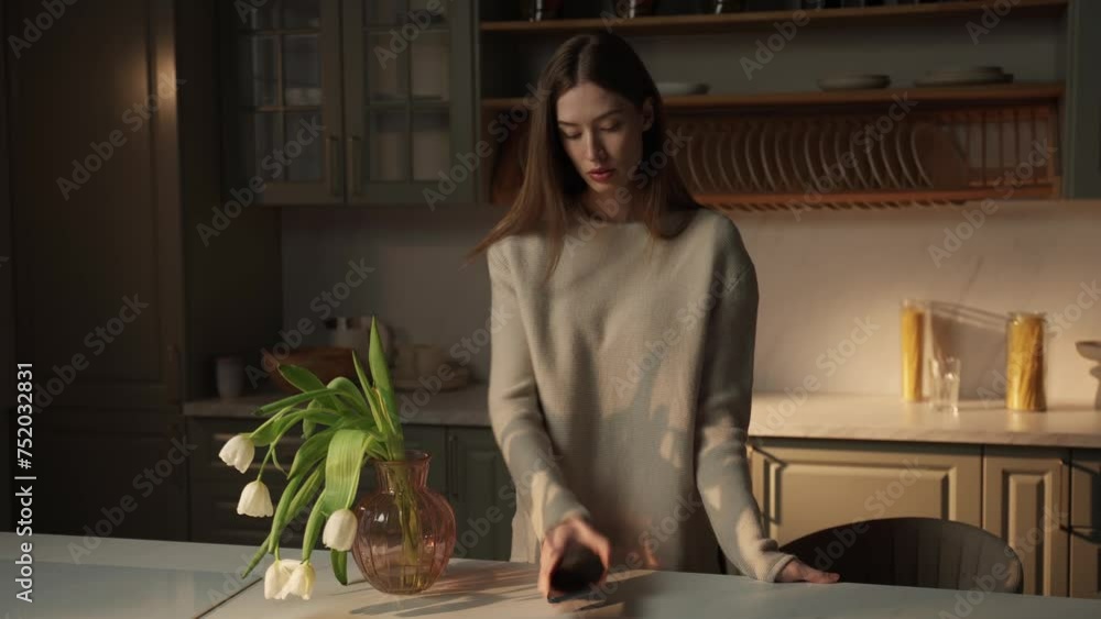 A young woman stands by a kitchen island, engrossed in her smartphone, with soft evening light filtering through the room. A vase of fresh tulips adds a touch of natural beauty to the tranquil A young woman stands by a kitchen island, engrossed in her smartphone, with soft evening light filtering through the room. A vase of fresh tulips adds a touch of natural beauty to the tranquil