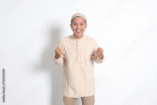 Portrait of religious Asian muslim man in koko shirt with skullcap raising his fist, celebrating success. Achievement concept. Isolated image on white background