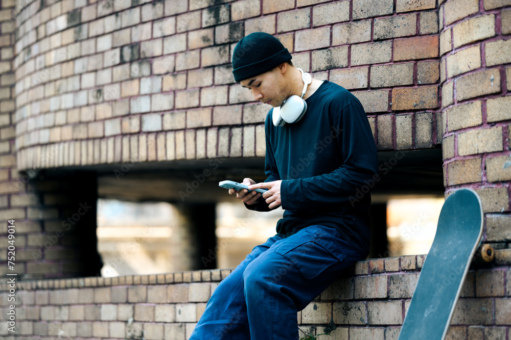 Handsome Asian man in stylish clothes using mobile phone while sitting with skateboard at city street Handsome Asian man in stylish clothes using mobile phone while sitting with skateboard at city street
