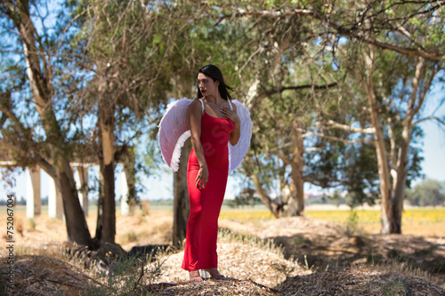 A young and beautiful brunette latin woman dressed in red with an elegant dress and white wings is doing different poses among eucalyptus trees. The woman is sad and desolate in the forest.