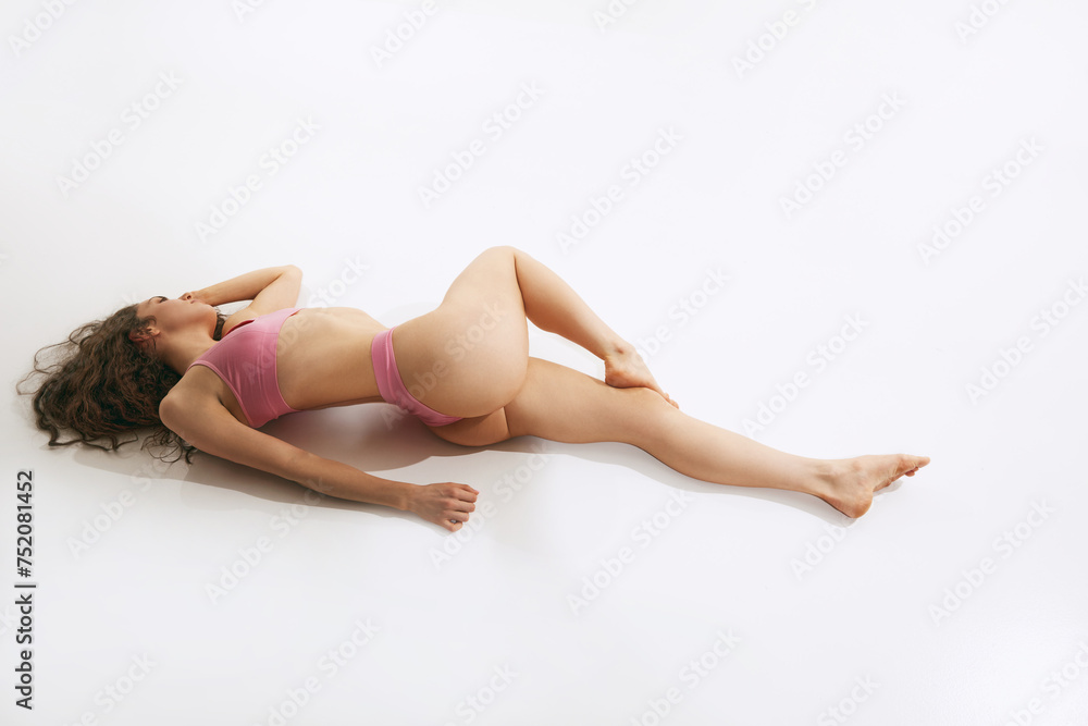 Full length portrait of young woman lying on floor and posing in lingerie against white studio background. Concept of beauty, style, fashion, cosmetology, body and skin care, sport, ad