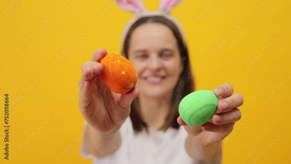 Easter joy and festivities. Springtime parties and events. Joyful delighted brown haired woman wearing bunny ears headband showing colored easter eggs posing isolated over yellow background Easter joy and festivities. Springtime parties and events. Joyful delighted brown haired woman wearing bunny ears headband showing colored easter eggs posing isolated over yellow background