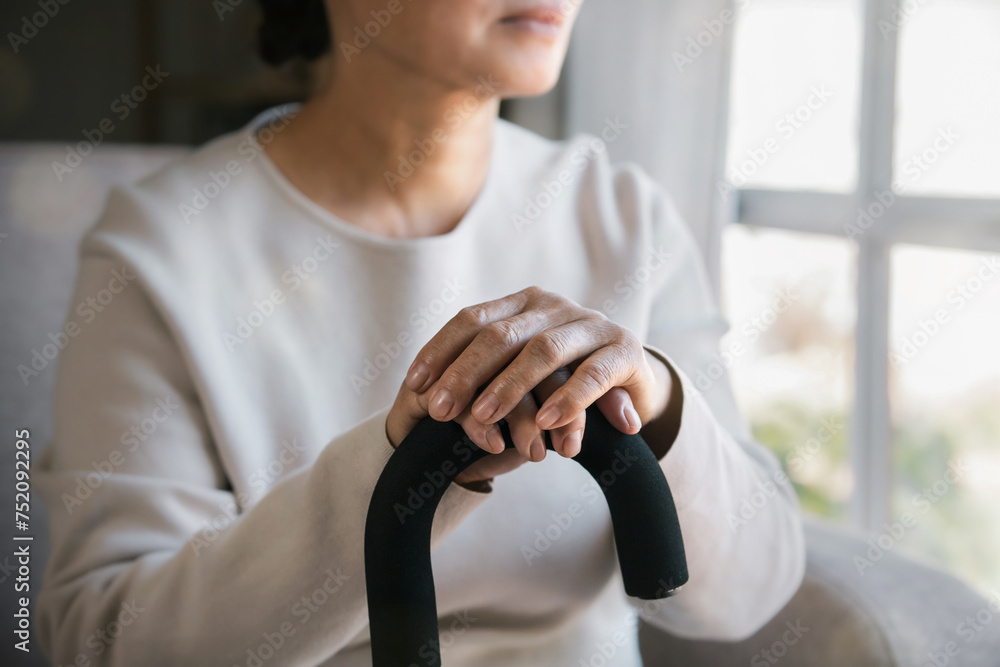 An elderly woman holding a walking stick sat alone and sad looking out the window. Think about health problems, loneliness, bad news, loss, suffering from health issues, sadness.