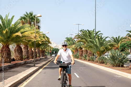 Happy retired senior man with helmet enjoying sport activity running with his electric bicycle in a sunny city street - palm trees on background - sustainable mobility concept