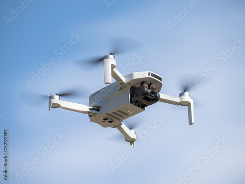 Bottom view of a small white drone with camera flying with its propellers out of focus due to speed flying through the air and a blue sky with clouds in the background
