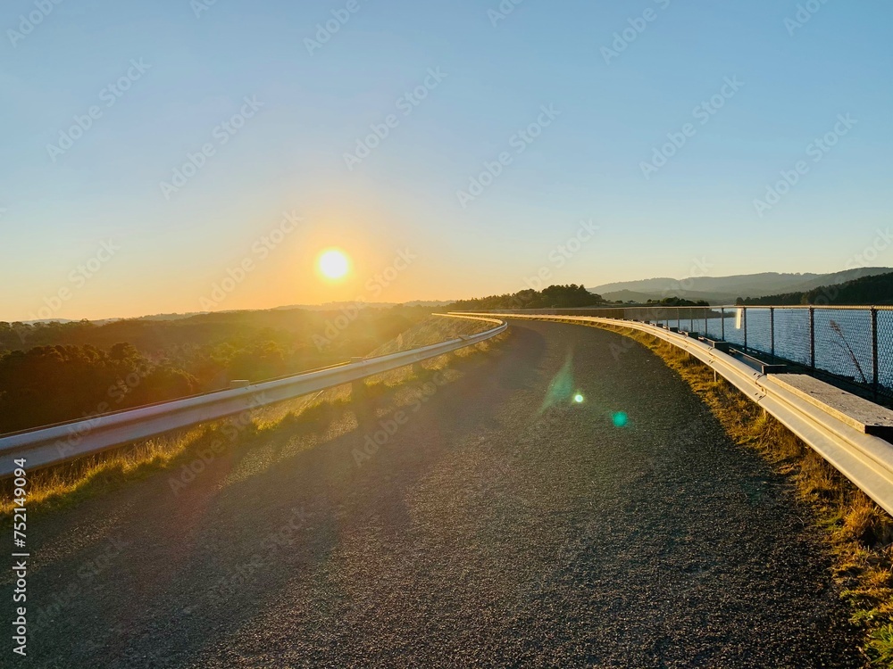Fototapeta premium A road with a railing alongside a beautiful forest field and a serene waterscape during sunset