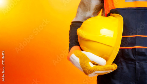 Manual worker with protective work gloves holding a yellow safety helmet on a yellow and orange background with copy space. Concept of workplace security. Generative Ai. © Alberto Masnovo
