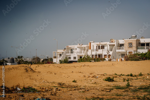 Agadir, Morocco - February 25, 2024 - Row of white houses behind a sandy open space with litter and sparse vegetation under a blue sky... © Raivo