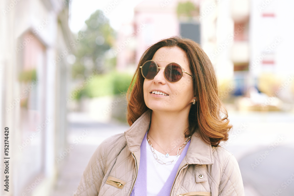 Outdoor portrait of happy middle age woman walking outside, wearing beige quilted jacket and sunglasses, nice sunny day - obrazy, fototapety, plakaty 