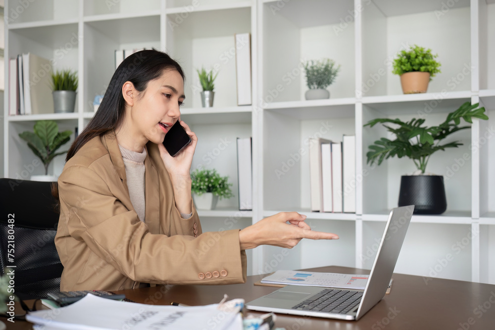 Young Asian business woman sits on the phone in an online business meeting using a laptop in a modern home office decorated with shady green plants.
