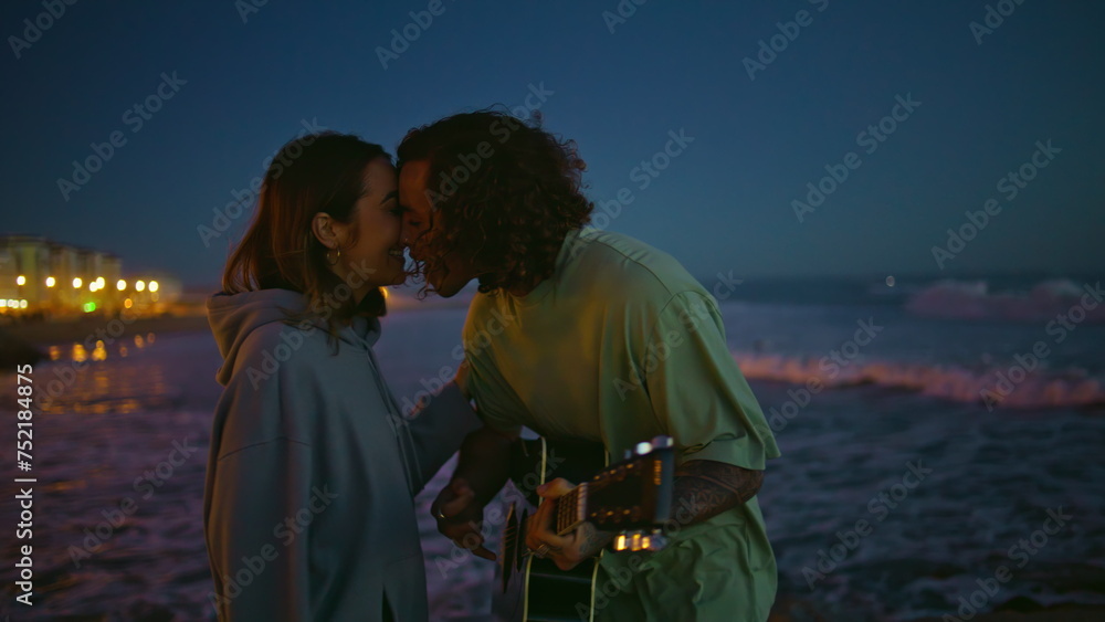 Fototapeta premium Enamoured man playing guitar at night sea beach closeup. Guy kissing girlfriend