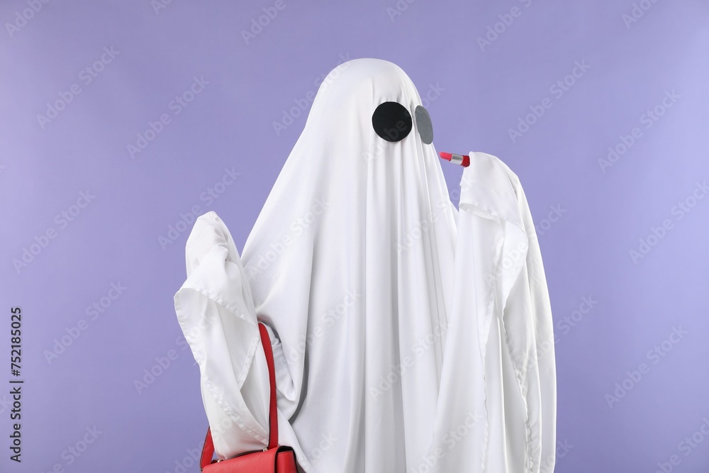 Glamorous ghost. Woman in white sheet with stylish bag and lipstick on violet background