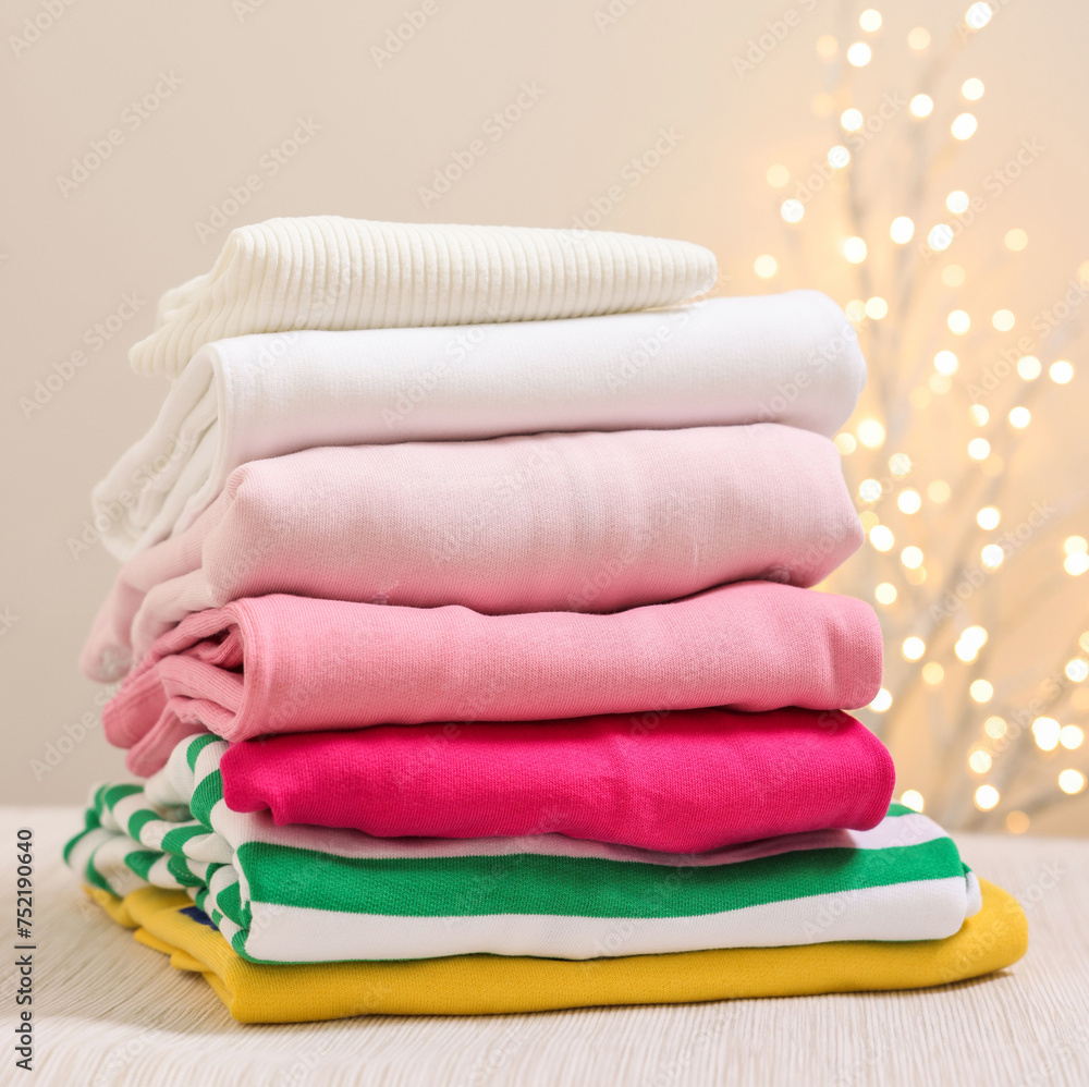 Stack of folded clothes on wooden table against beige background with blurred lights, space for text Stack of folded clothes on wooden table against beige background with blurred lights, space for text