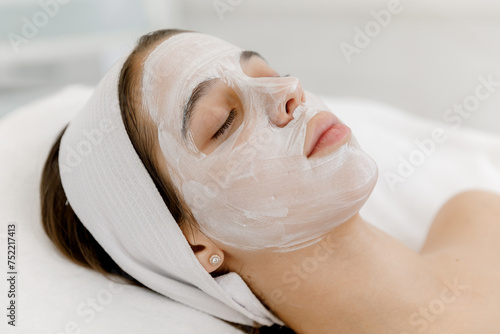 A teenage girl with a white mask on her face is lying on a cosmetologist’s couch. Cosmetology and facial skin care in beauty salon. White mask. 