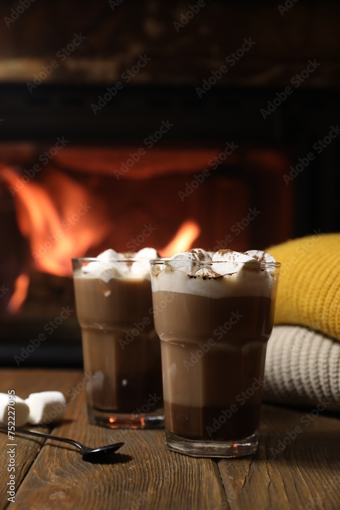 Glasses with hot cocoa, marshmallows and sweaters on wooden table near fireplace