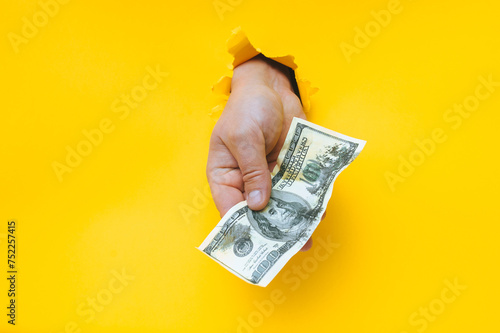 A man's hand holds dirty money through a torn hole in yellow paper. Concept of dishonest income, donation, profit and salary fraud.