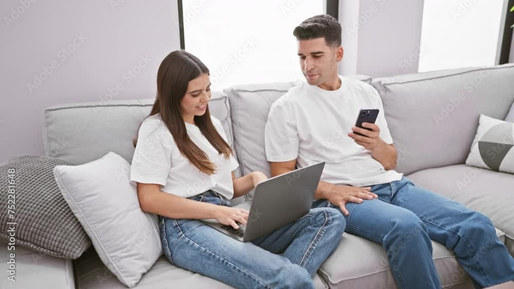 A man and a woman wearing casual clothes while sitting on a gray sofa in a well-lit living room, the man holds a smartphone and the woman uses a laptop. A man and a woman wearing casual clothes while sitting on a gray sofa in a well-lit living room, the man holds a smartphone and the woman uses a laptop.