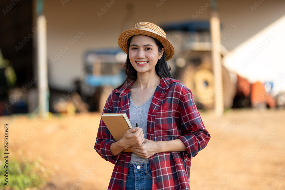 Proud female farmer stands watching and taking notes on the growth of her farm with a tractor behind her.