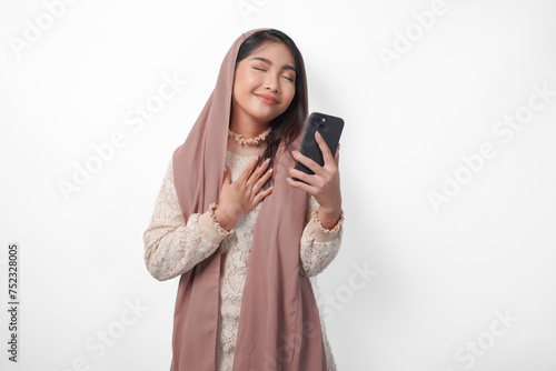 Happy mindful thankful young Asian Muslim woman hand on chest smiling while holding smartphone, isolated on white background