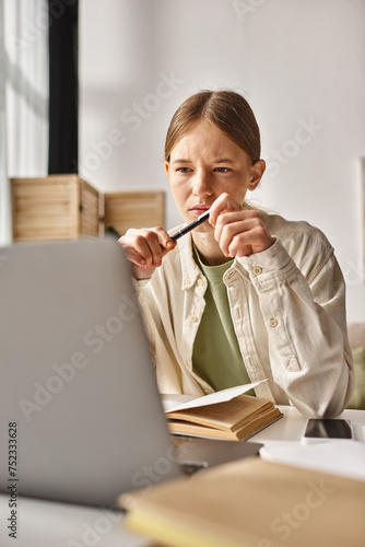 teenage girl holding pen and looking at laptop while doing her homework near book and phone on desk
