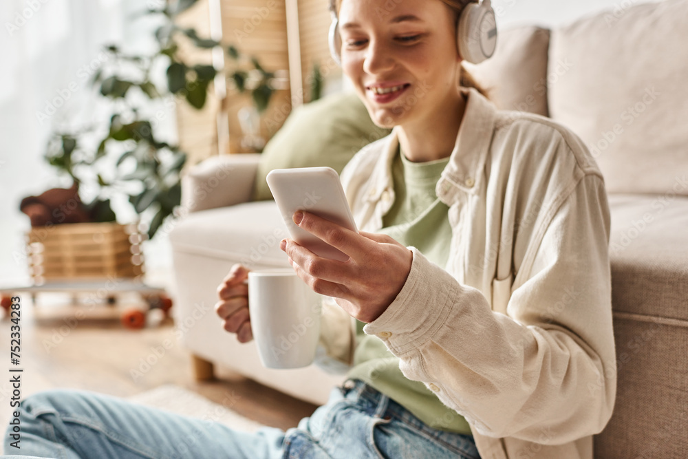 smiling teenager girl in wireless headphones using smartphone while holding cup of tea at home