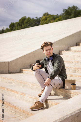 A teenager in headphones with a phone in his hands sits on the steps in the street