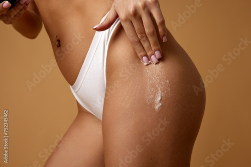 Close up of woman applying scrubbing product on legs against sandy color studio background. Orange-peel skin prevention. Concept of anti-cellulite, dermatology treatments, cosmetology care.