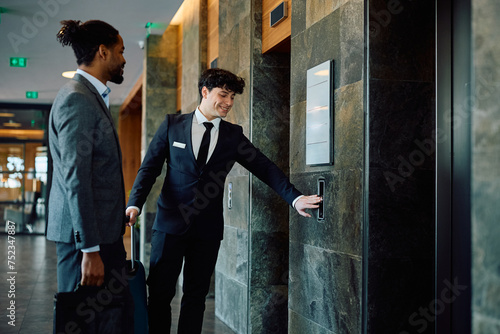 Happy hotel attendant and black businessman waiting for elevator in lobby, photo