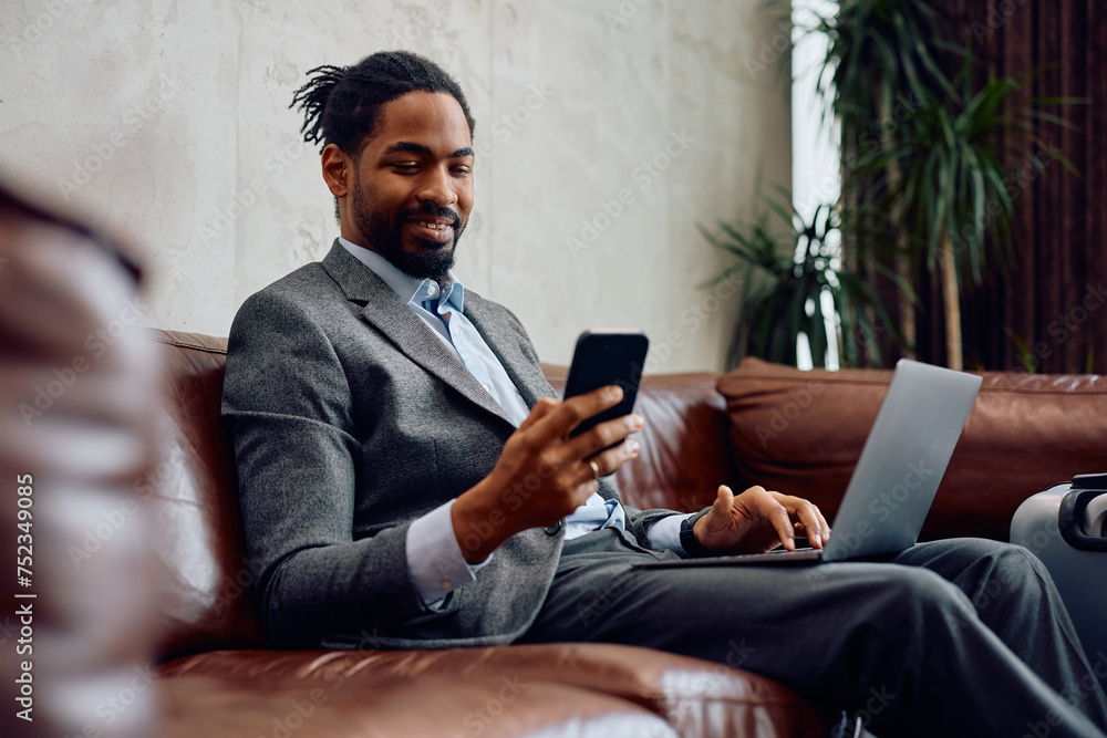 Happy black businessman texting on cell phone while working on laptop in hotel lobby. Happy black businessman texting on cell phone while working on laptop in hotel lobby.