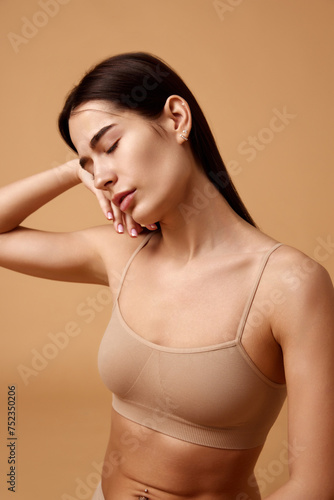 Young beautiful sensual brunette with closed eyes posing in beige lingerie against sandy color studio background. Concept of beauty, spa procedures, dermatology treatments, cosmetology care.