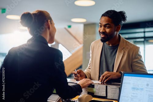 Happy African American man talking to hotel receptionist while filing registration paperwork at front desk. photo