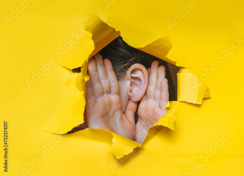 Female ear and hands close-up. Copy space. Torn paper, yellow background. The concept of eavesdropping, espionage, gossip and the yellow press.