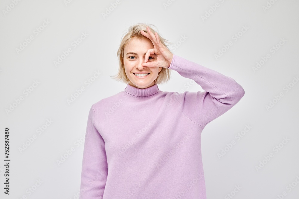 Smiling woman in purple turtleneck making OK gesture with hand over eye on light background Smiling woman in purple turtleneck making OK gesture with hand over eye on light background