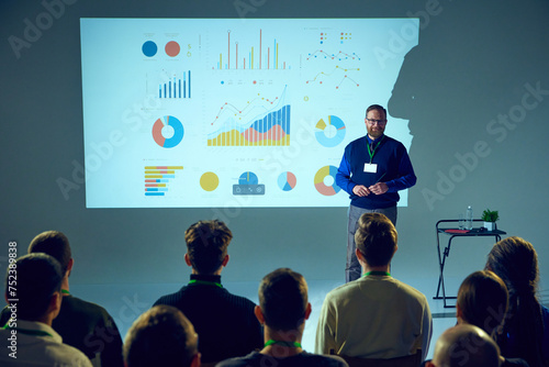 Presenter with ID badge stands in front of an audience, giving lecture with data chart backdrop in conference room. Concept of business, startup, leadership and personal development courses. © Lustre