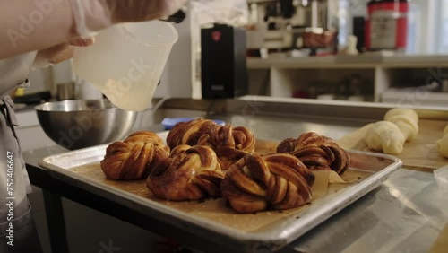 The baker uses a brush to apply syrup to freshly baked, hot cinnamon rolls.